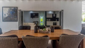 A dining area with a wooden table and wicker chairs, a painting on the wall, and a kitchen in the background. - Palm Beach 34 Suitable for 8 Persons 4 Bedrooms 4 Bathrooms (Noord)