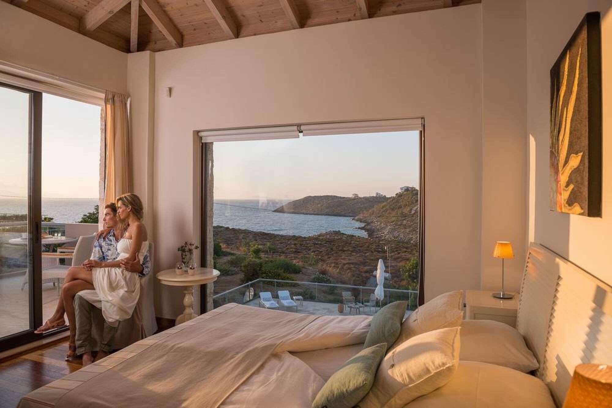 A bedroom with a large window overlooking a coastal landscape, a bed, a small table, and a lamp.