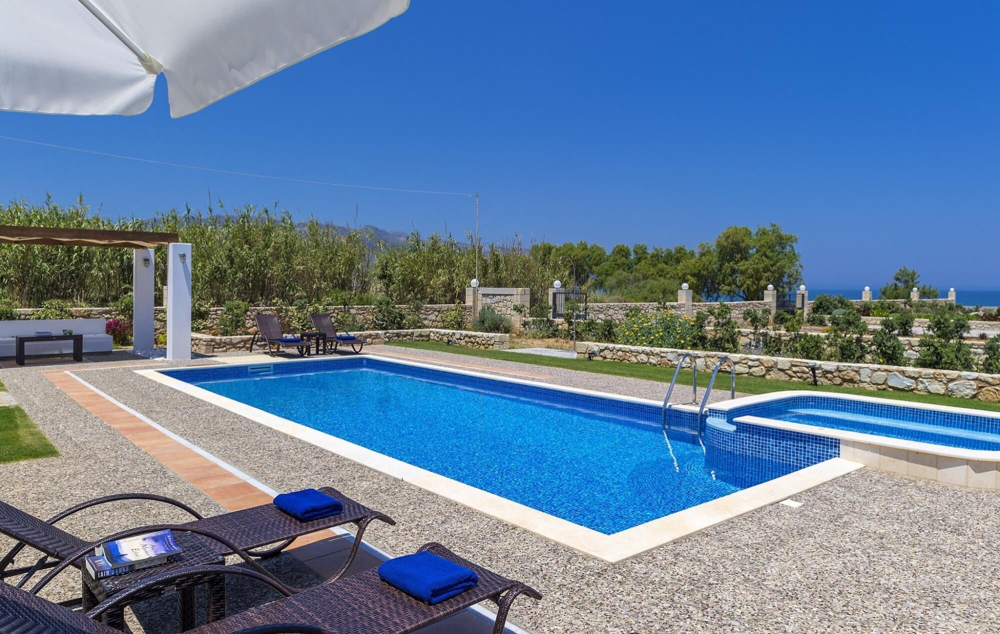 Una piscina rectangular al aire libre con agua azul cristalina, rodeada por un muro de piedra y una zona ajardinada. Hay sillones de descanso y una pérgola con una sombrilla blanca.