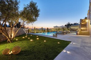 A pool area with a stone wall, a grassy lawn, and a pergola with string lights. - Villa Rouga in Xirosterni (Apokoronas)
