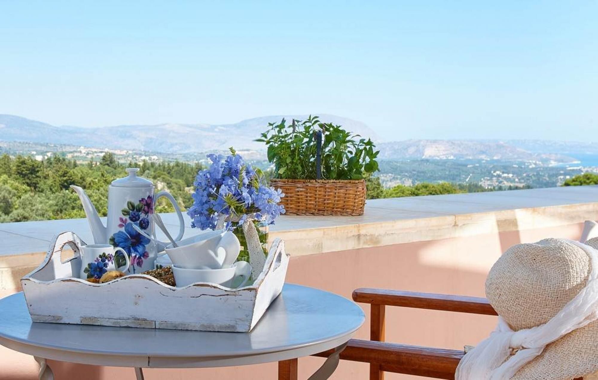 A table with a teapot, cups, and a floral arrangement on a terrace with a view of mountains and greenery.