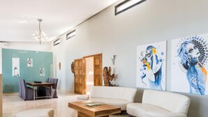 A spacious living room with a white sofa, a wooden coffee table, and a dining area with a table and chairs. There are two large paintings on the wall and a wooden door. - Jamila Villa in Kalathas (Chania)