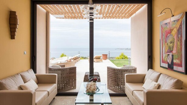 A modern living room with a glass coffee table, two beige sofas, and a view of the sea through large windows. - Jamila Villa in Kalathas (Chania)
