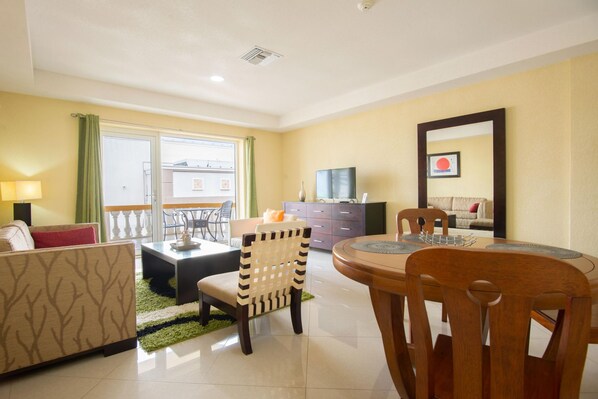 A living room with a sofa, a dining table with chairs, a TV, and a balcony with a view. - Bay Harbor Luxury 1 Bedroom Apartments (Oranjestad)