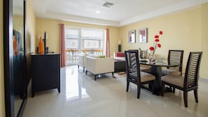 A dining area with a table set for four, surrounded by chairs. There is a sofa and a small table with a lamp in the background.
