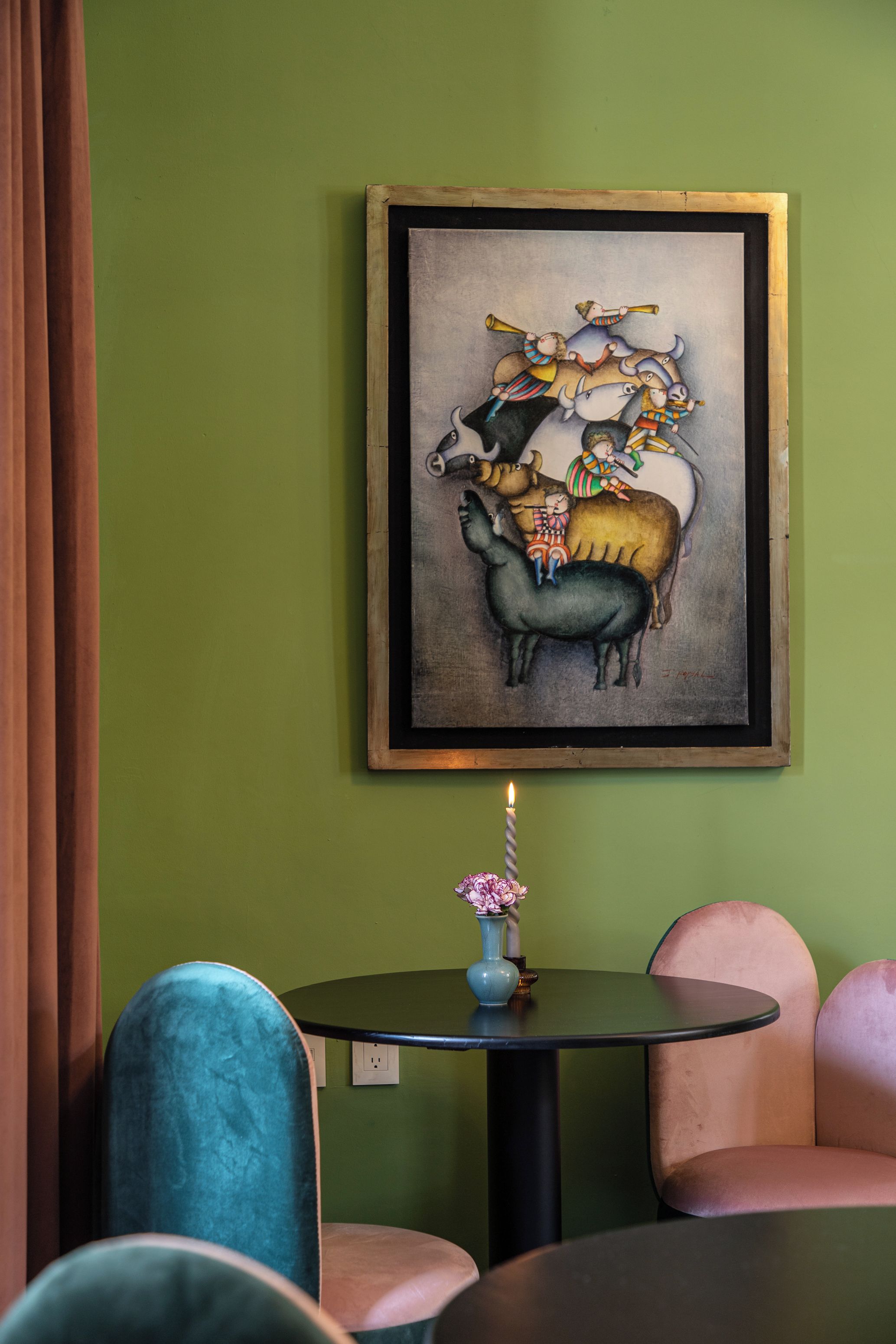 A dining area with a green wall, a framed painting of animals, a small table with a vase, and teal and pink chairs.