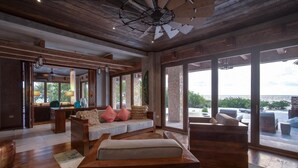A spacious living area with a large window offering a view of the ocean, a wooden ceiling fan, and a cozy seating arrangement. - Gladden Caye Private Island Resort (Placencia)