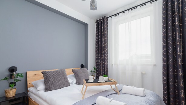 Una habitación de hotel moderna con una cama de madera, ropa de cama gris, una mesita de noche con lámpara y una ventana con cortinas.
