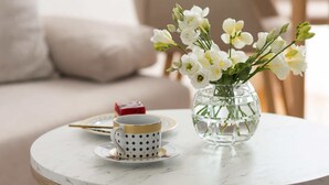 A round wooden table with a clear glass vase containing white flowers, a white cup with a gold rim, and a red box on a white marble surface.
