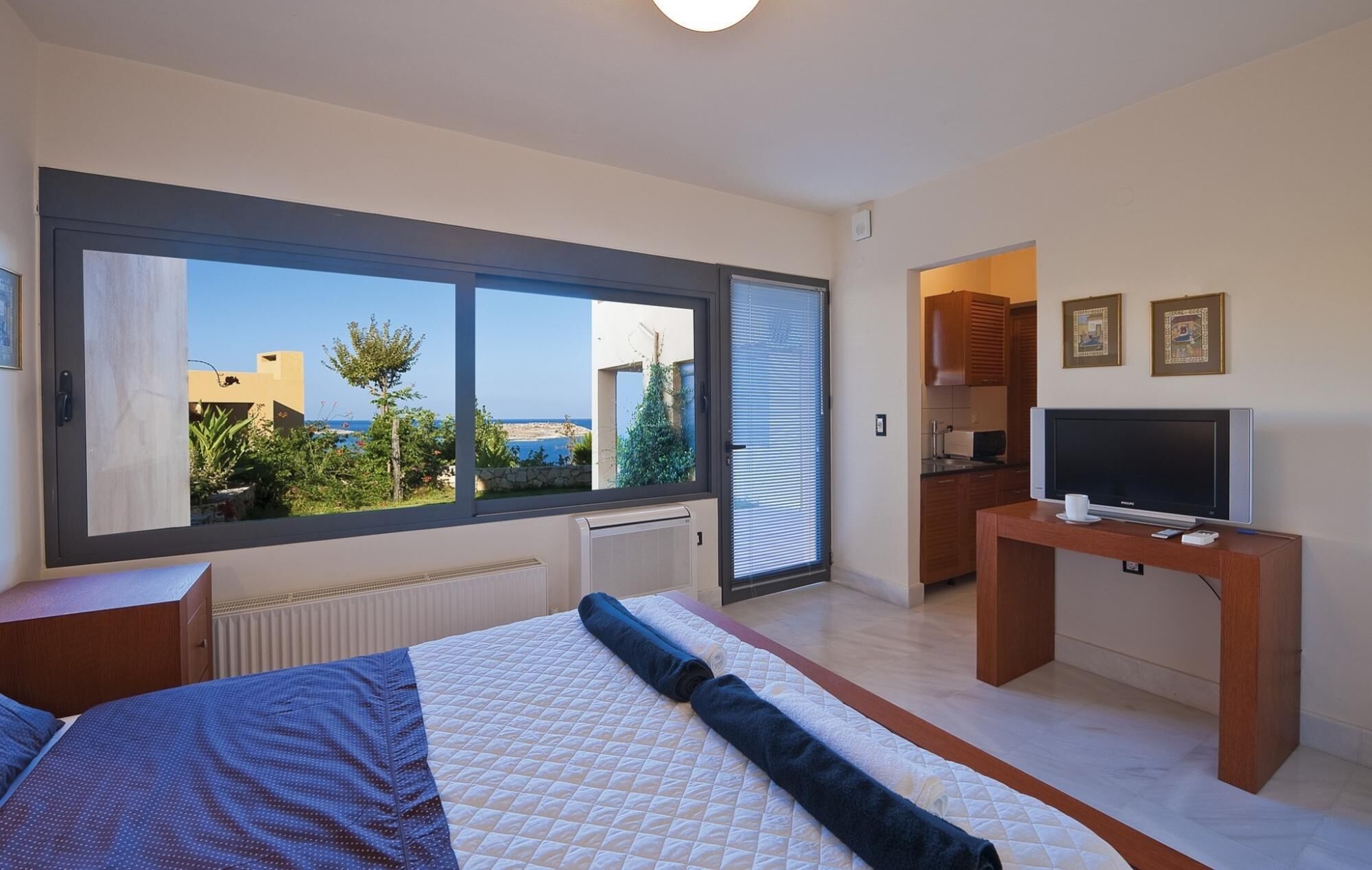 A bedroom with a large window, a bed, a TV, and a kitchenette.