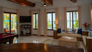 A spacious living room with a pool table, comfortable seating, large windows, and a ceiling fan. - Villa Island Time Gecko (Placencia)
