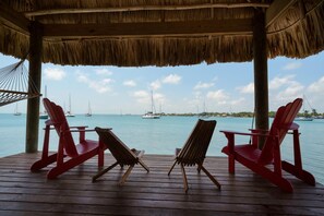 Eine Holzterrasse mit zwei roten Adirondack-Stühlen und einer Hängematte, mit Blick auf ruhiges Wasser und Segelboote.