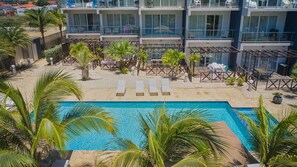 Área de piscina com espreguiçadeiras e palmeiras, cercada por um edifício com várias varandas e assentos externos.