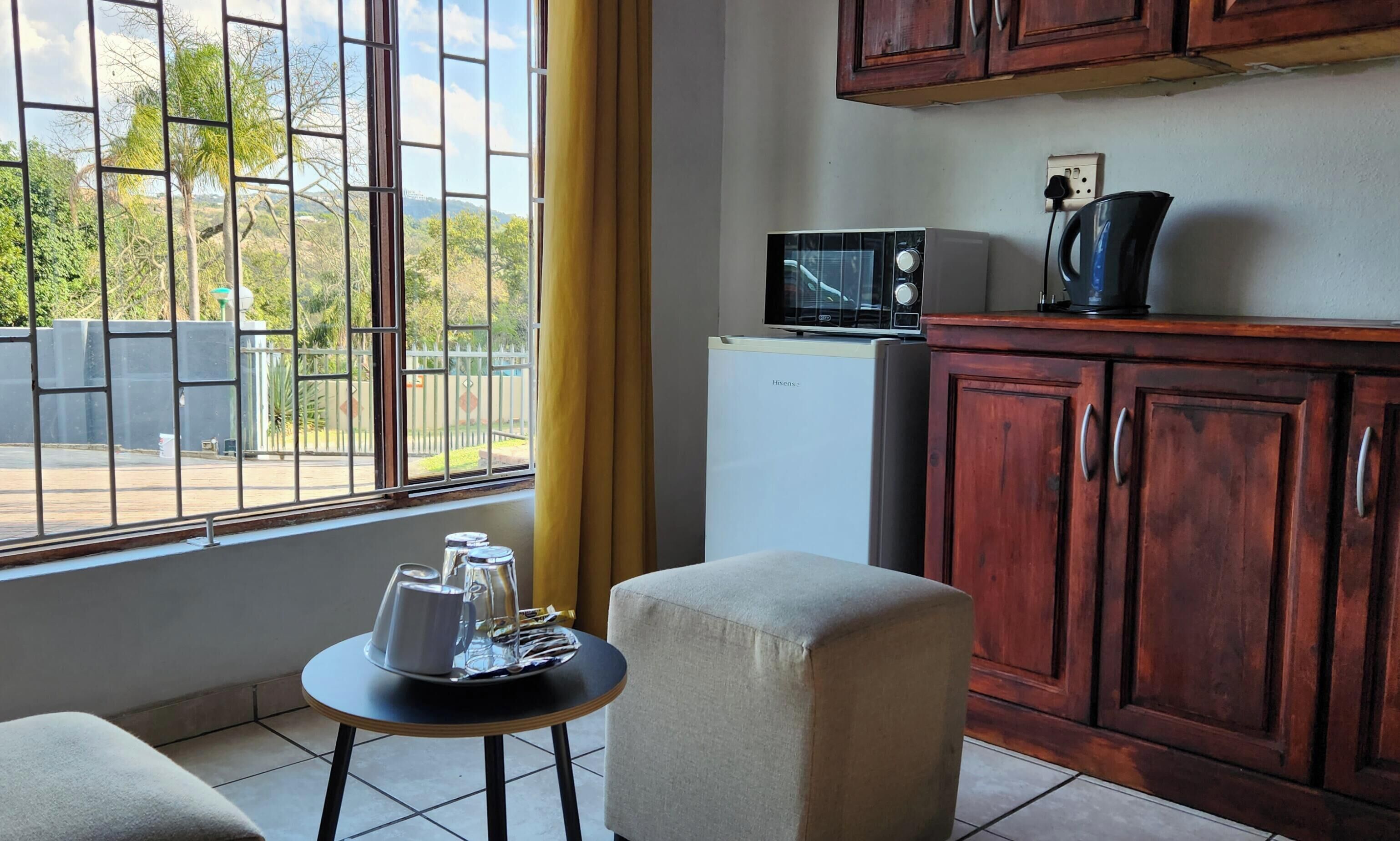 A kitchen with a microwave, refrigerator, and wooden cabinets. There is a small table with a pitcher and glasses, and a window with a view of greenery and a fence.