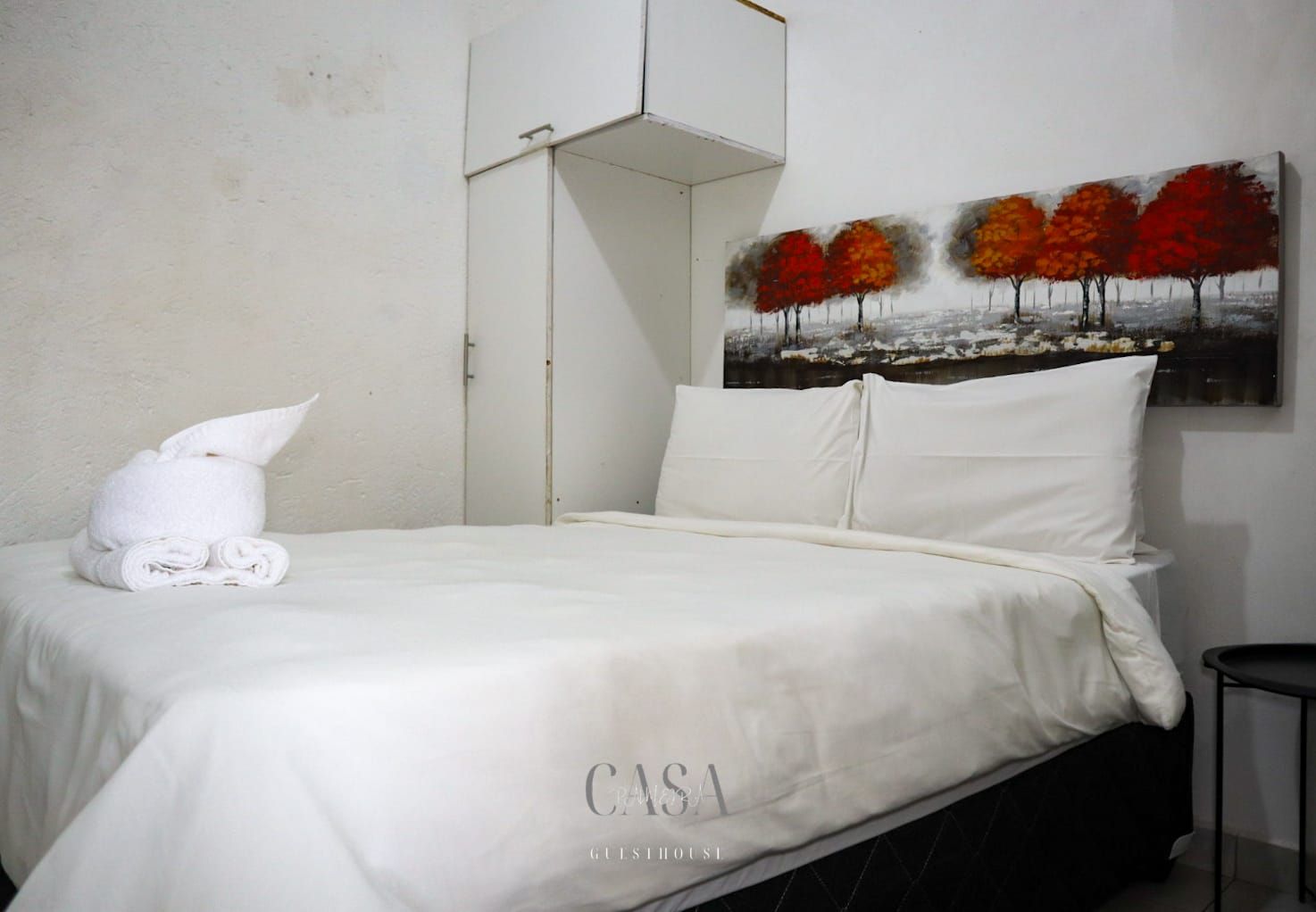 A hotel room with a neatly made bed, a painting of autumn trees on the wall, and a folded towel on the bed.