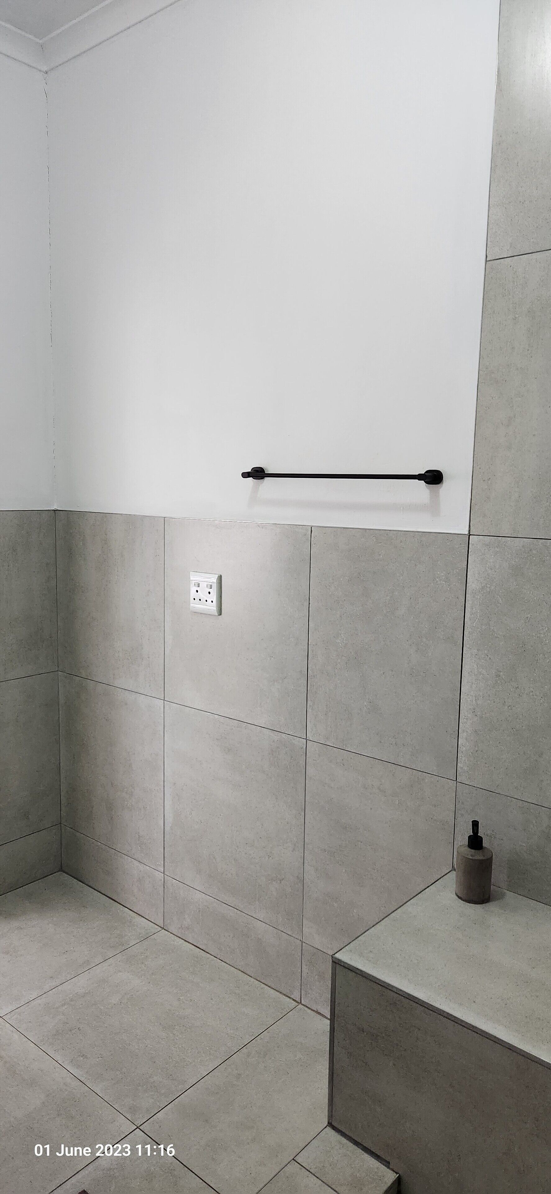 A bathroom with grey tiled walls and floor, a black towel rack, a white electrical outlet, and a small bottle on a shelf.