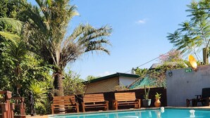 Un espace piscine doté d’une terrasse carrelée, de chaises longues en bois et d’un palmier.