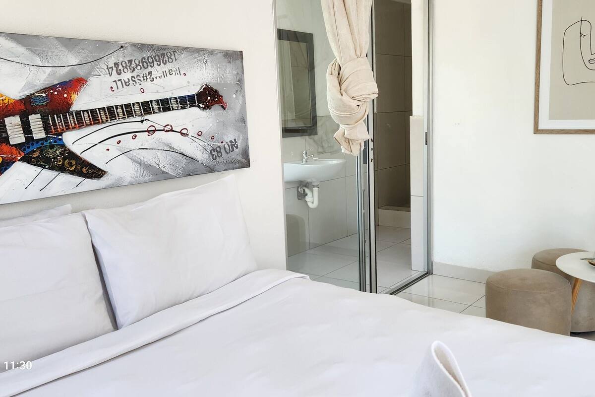 Habitación de hotel con una cama, un cuadro de una guitarra, un baño con ducha, una mesita con hervidor y un marco con una fotografía en la pared.