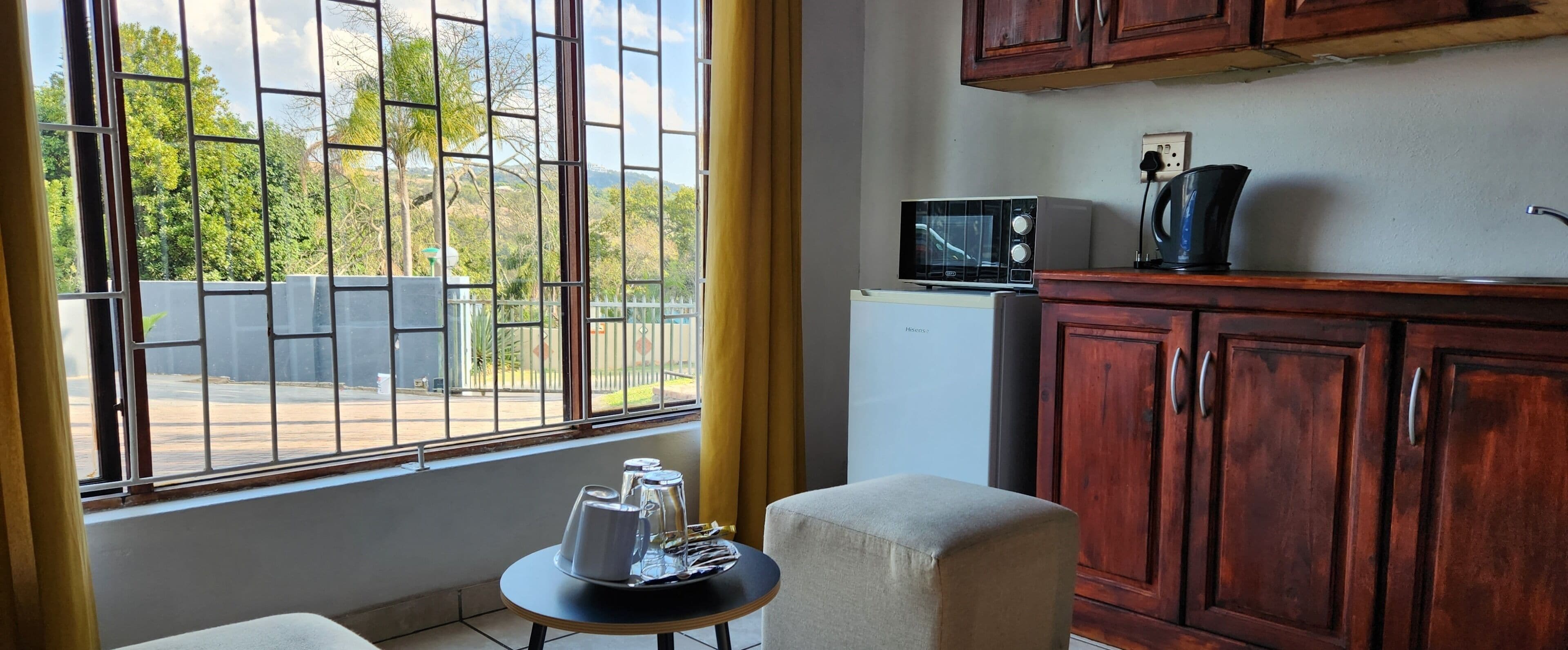 A kitchen with a microwave, refrigerator, and wooden cabinets. There is a small table with a pitcher and glasses, and a window with a view of greenery and a fence.