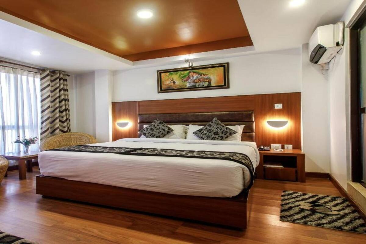 A hotel room with a large bed, a wicker chair, a small table with a vase, and a painting on the wall.