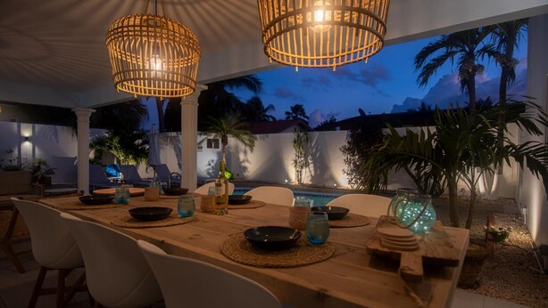 An outdoor dining area with a wooden table, white chairs, and two wicker pendant lights. - NEW Cozy Casa in Oranjestad (Oranjestad)