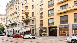 A street view with a row of parked cars, a tram track, and buildings with shops on the ground floor.