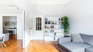 A modern living room with a grey sofa, a white bookshelf, and a dining area with a white table and chairs.