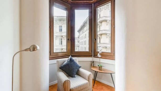 A corner room with a striped armchair, a small table with a plant, and two windows offering a view of buildings.