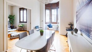 A modern living room with a large white table, comfortable seating, a decorative ceiling light, and a view of the city through large windows.