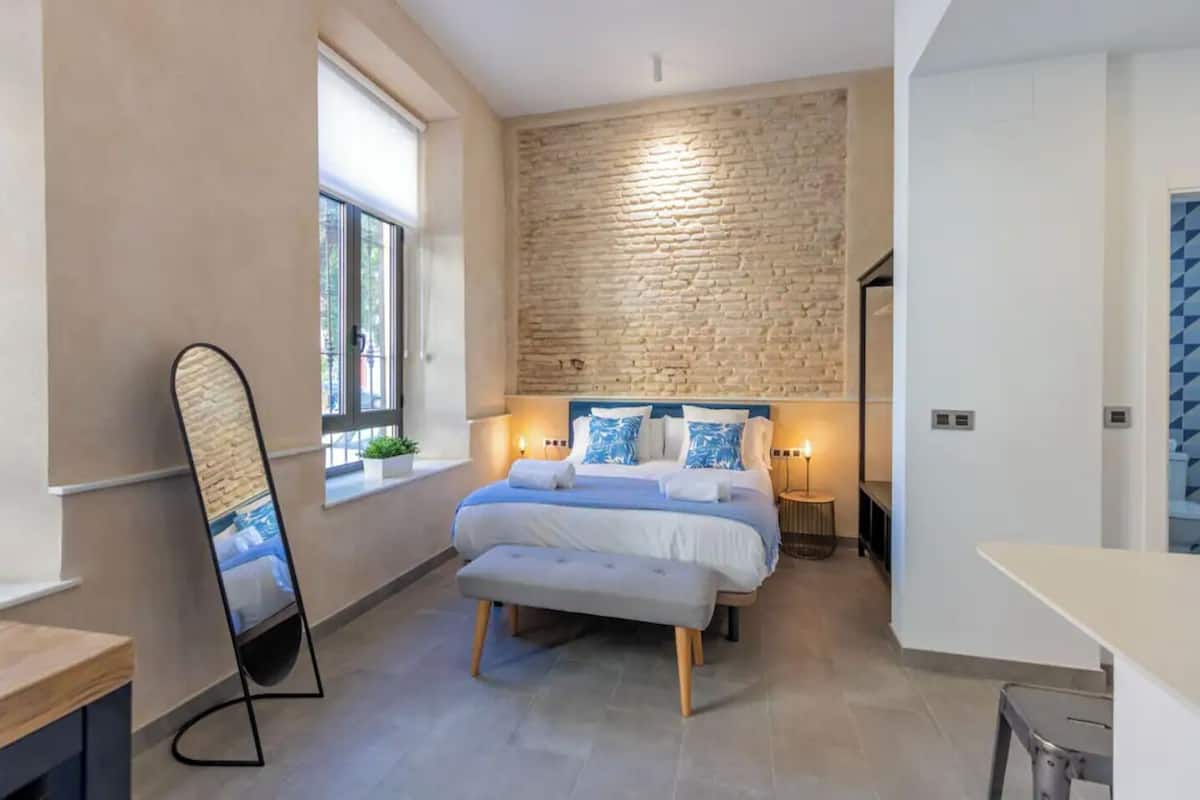 A modern bedroom with a large bed, a brick accent wall, a bedside table with a lamp, and a window with a view.