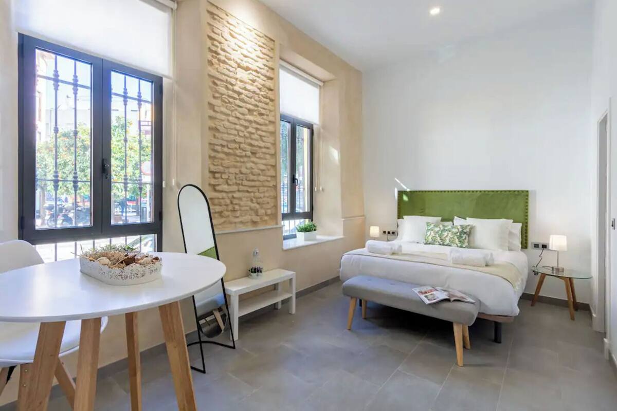 A modern hotel room with a bed, bedside tables, a round dining table, and a stone accent wall.