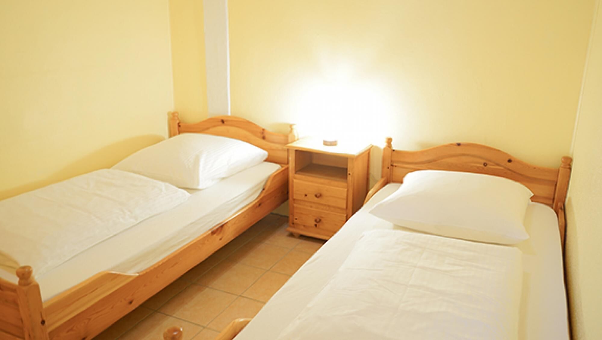 Dos camas individuales de madera con ropa blanca, una mesita de noche con cajones y una lámpara fijada a la pared.