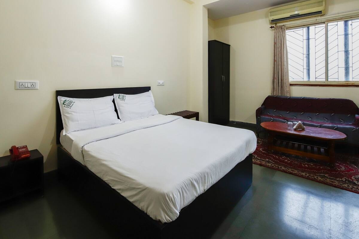 A hotel room with a bed, a small table, a sofa, an air conditioner, and a window with curtains.