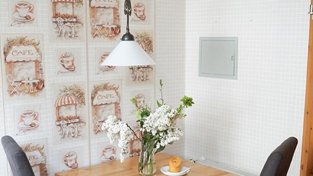 A small dining area with a wooden table and two chairs, a hanging lamp, and a wall with a cafe-themed wallpaper.