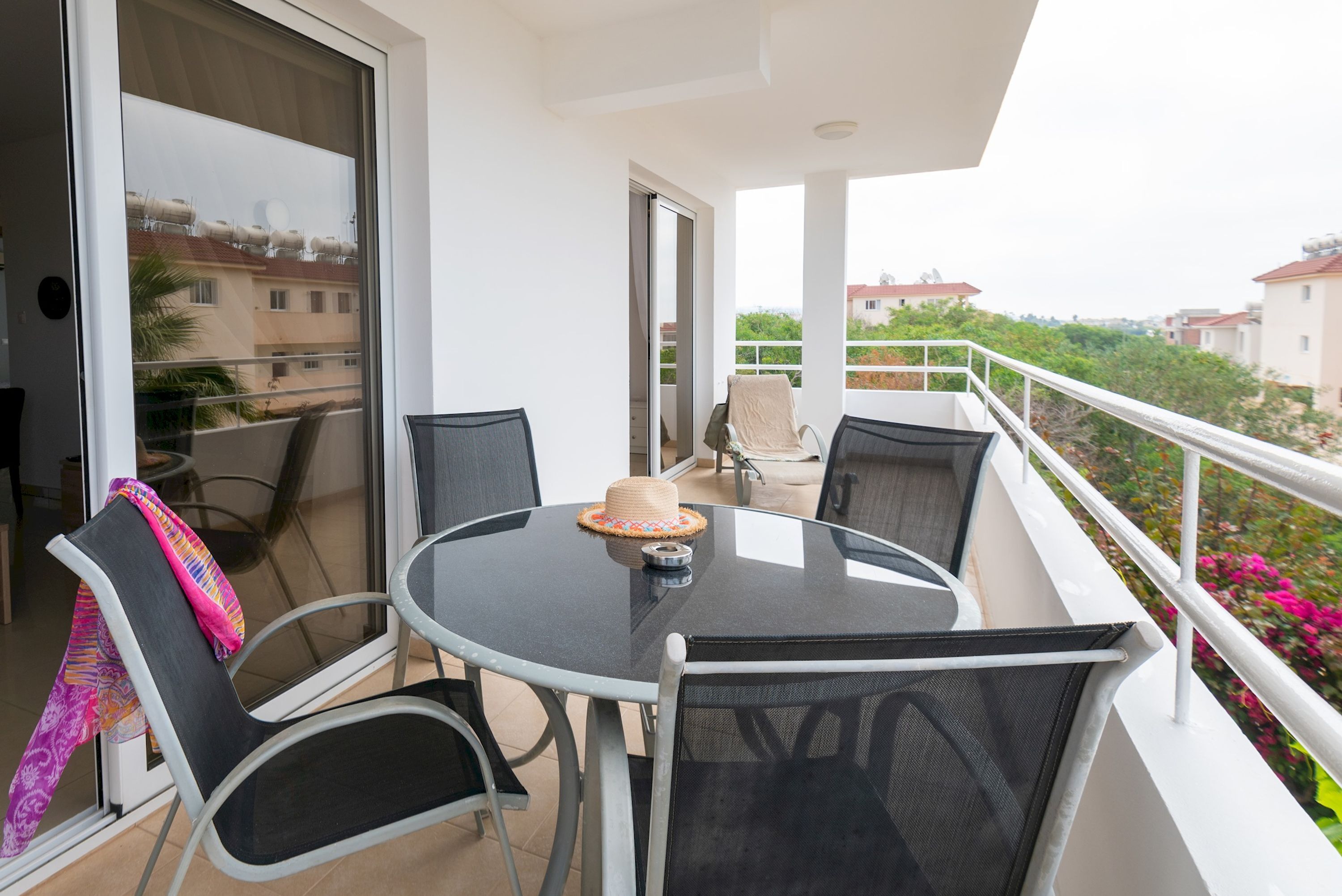 A balcony with a round table and four chairs, a sliding glass door, and a view of neighboring buildings and greenery.