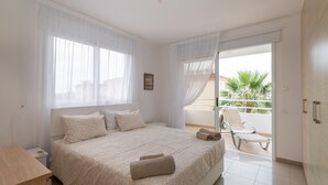 A bedroom with a large bed, a nightstand, a wooden side table, and a balcony with a chair. - Narcissos Nissi Beach Apartment A8 (Ayia Napa)