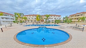 A swimming pool with a tiled deck, surrounded by palm trees and buildings with red roofs. - Narcissos Nissi Beach Apartment A8 (Ayia Napa)
