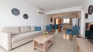 A modern living room with a sofa, coffee table, and chairs. There is a kitchen in the background with a refrigerator and a microwave. - Narcissos Nissi Beach Apartment A8 (Ayia Napa)