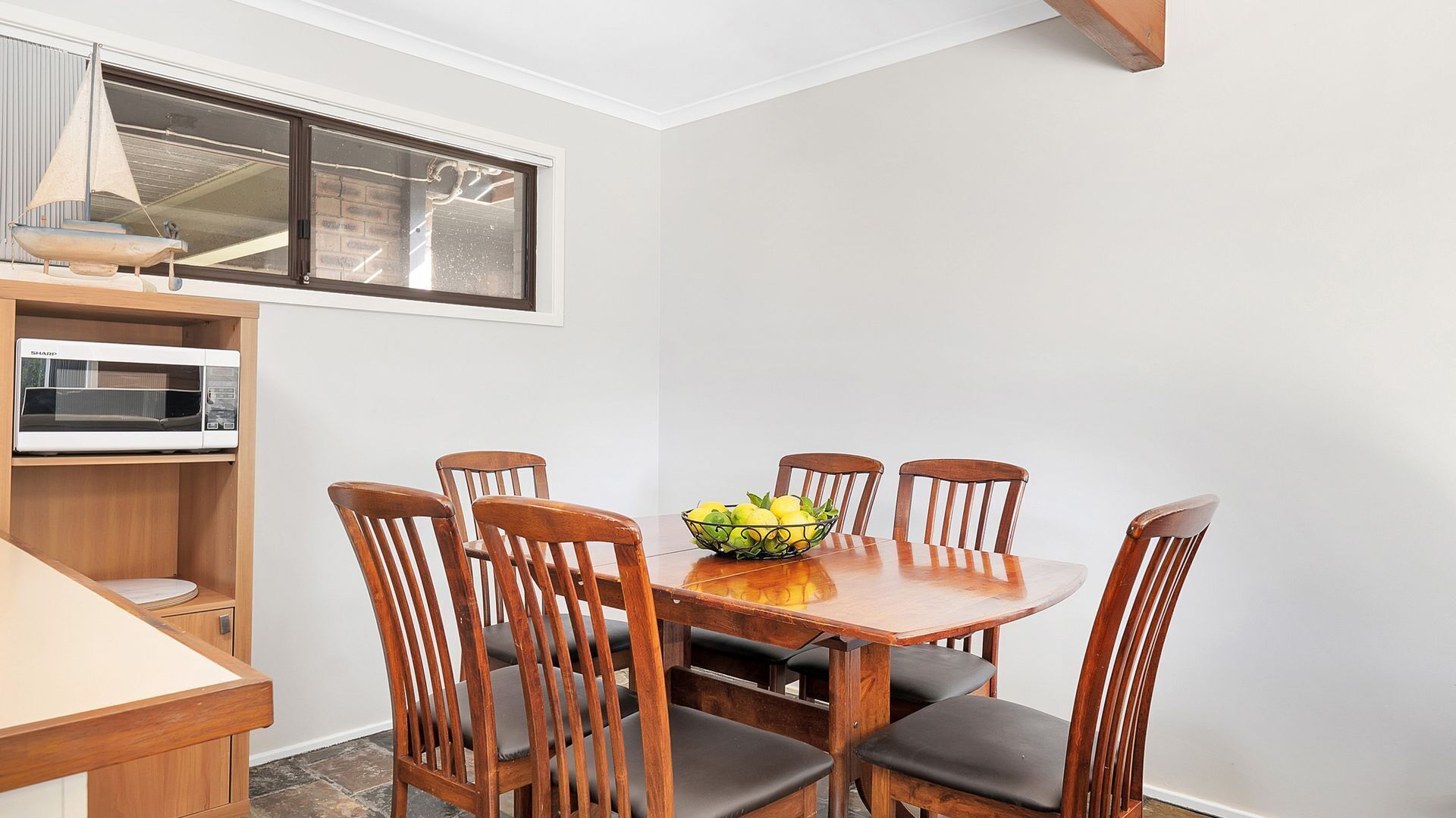Un coin repas avec une table en bois et six chaises, un saladier de fruits sur la table, un four  à micro-ondes sur une étagère et une fenêtre décorée avec un motif de voilier.