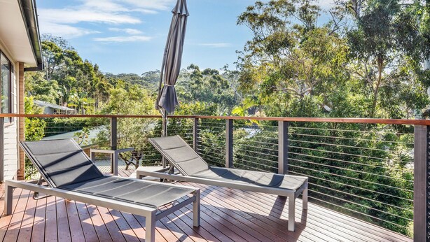 A balcony with two sun loungers, a metal railing, and a view of a forested area. - Fields of Green by Experience Jervis Bay (Vincentia)