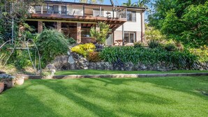 A modern two-story house with a stone retaining wall, a well-maintained lawn, and a variety of tropical plants. - Fields of Green by Experience Jervis Bay (Vincentia)