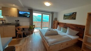 A cozy bedroom with a wooden bed, a desk with a chair, and a balcony with a view of mountains. - Studio in Ötztal with wellness, children's playground and farm (Umhausen)