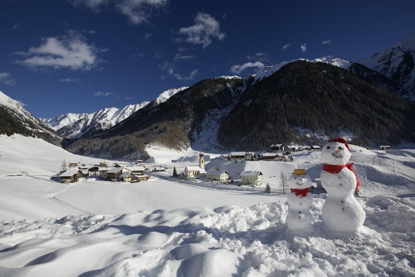 A snowman in a snowy landscape with a village and mountains in the background.