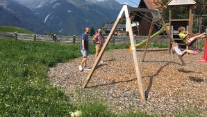A playground with a swing set and a slide, surrounded by a wooden fence and a view of mountains. - Studio in Ötztal with wellness, children's playground and farm (Umhausen)