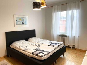 Ein Schlafzimmer mit Bett, Heizkörper, einem Fenster mit Vorhängen und einer Deckenleuchte.