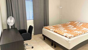 Une chambre à coucher comprenant un lit, un bureau, une chaise, un ventilateur sur pied et une fenêtre avec des rideaux.