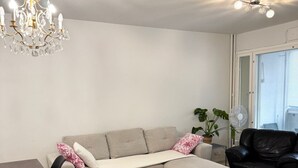 Un salon comprenant un canapé d’angle, un fauteuil en cuir noir, une table à manger, un lustre et une grande fenêtre avec des stores.