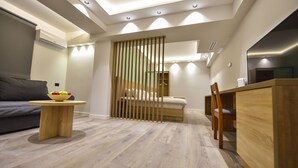 Una habitación moderna y bien iluminada con una cama, un escritorio de madera, una pequeña mesa redonda con un frutero y un sofá gris.