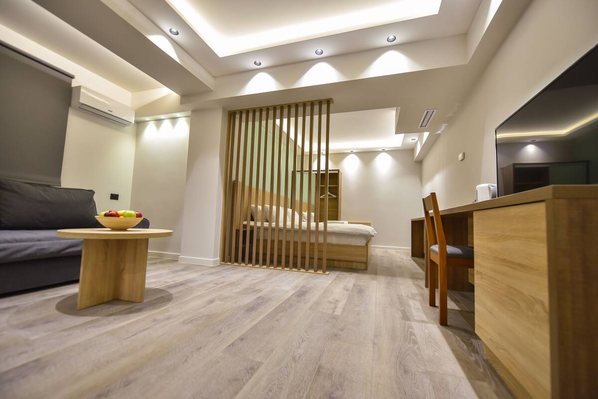 A modern, well-lit room with a bed, a wooden desk, a small round table with a bowl of fruit, and a grey sofa.