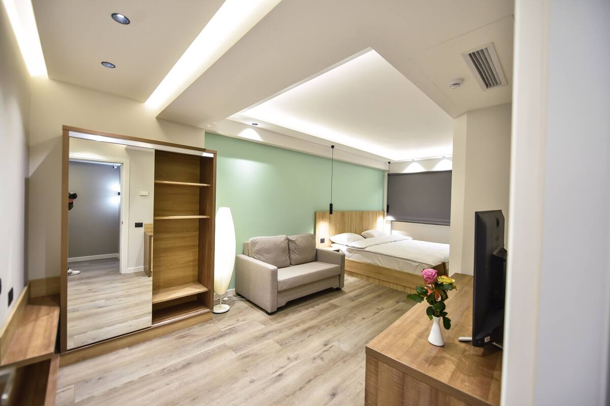 A modern hotel room with a wooden floor, a sofa, a bed, a TV, and a mirror.